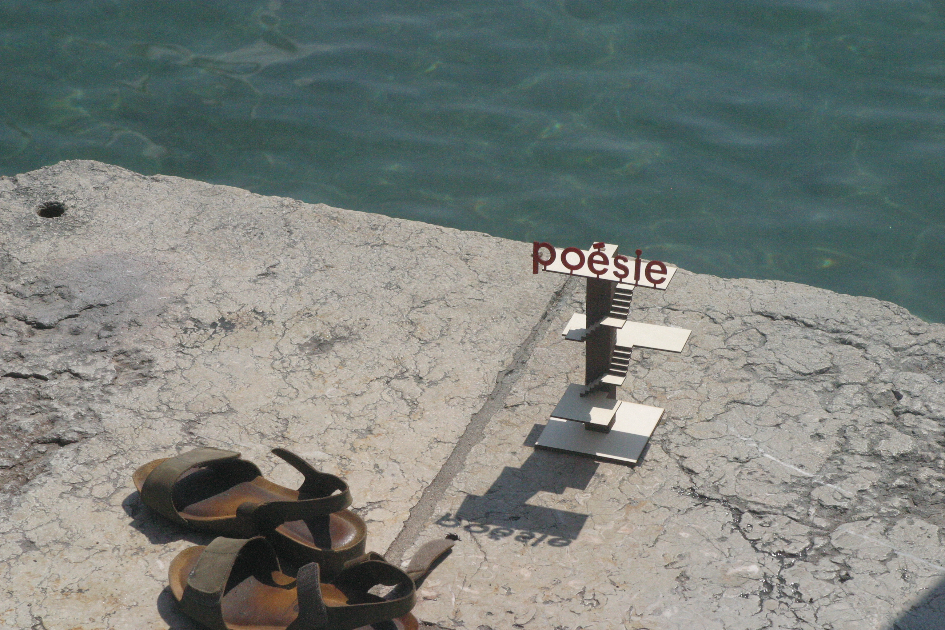 Maquette du plongeoir des Pâquis surmontée du mot « poésie », posée sur le quai au bord du lac, près d'une paire de sandales